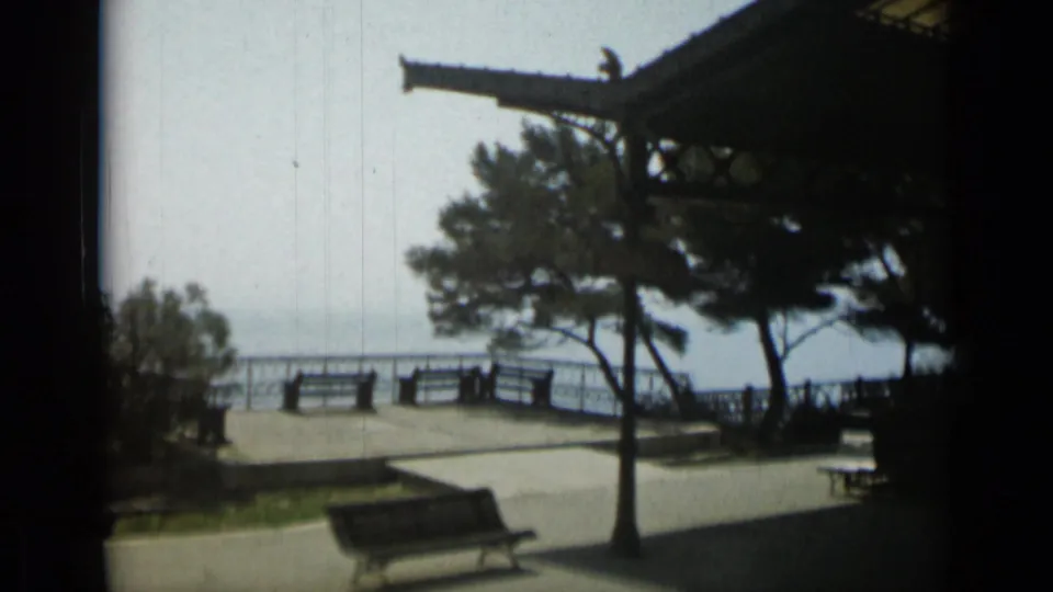 1962 Monaco seaside promenade with benches, railings, trees, open water beyond, and part of a covered pavilion structure overhead.