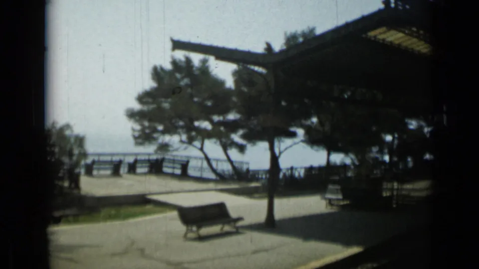 1962 home-movie view of a seaside rail platform with benches, pine trees, a canopy and water beyond the railing.