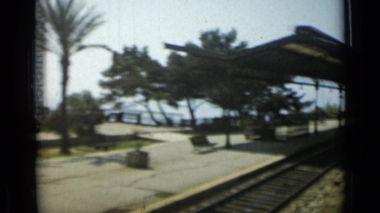 Rail tracks beside a platform with trees, benches, waterfront railing and station canopy.