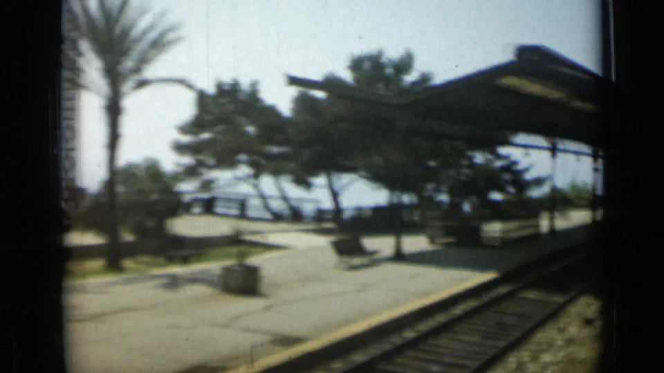 Moving-train view of a sunlit Riviera-style platform, rail tracks, benches, trees and water-facing railings.