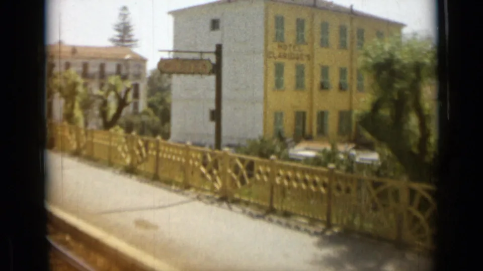 Passing train view of a yellow town building with signage, parked cars, a sidewalk and decorative fence near the tracks.