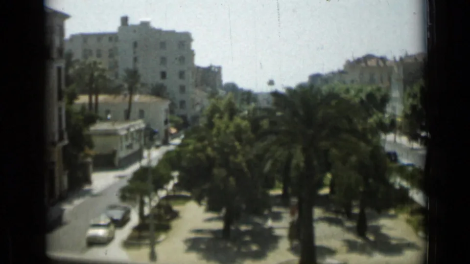 1962 Monaco view over palms and parked cars toward tall pale buildings, filmed from an elevated visitor perspective.