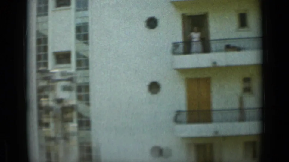 Monaco apartment facade with balconies and a visible person standing on an upper balcony in 1960s home-movie footage.