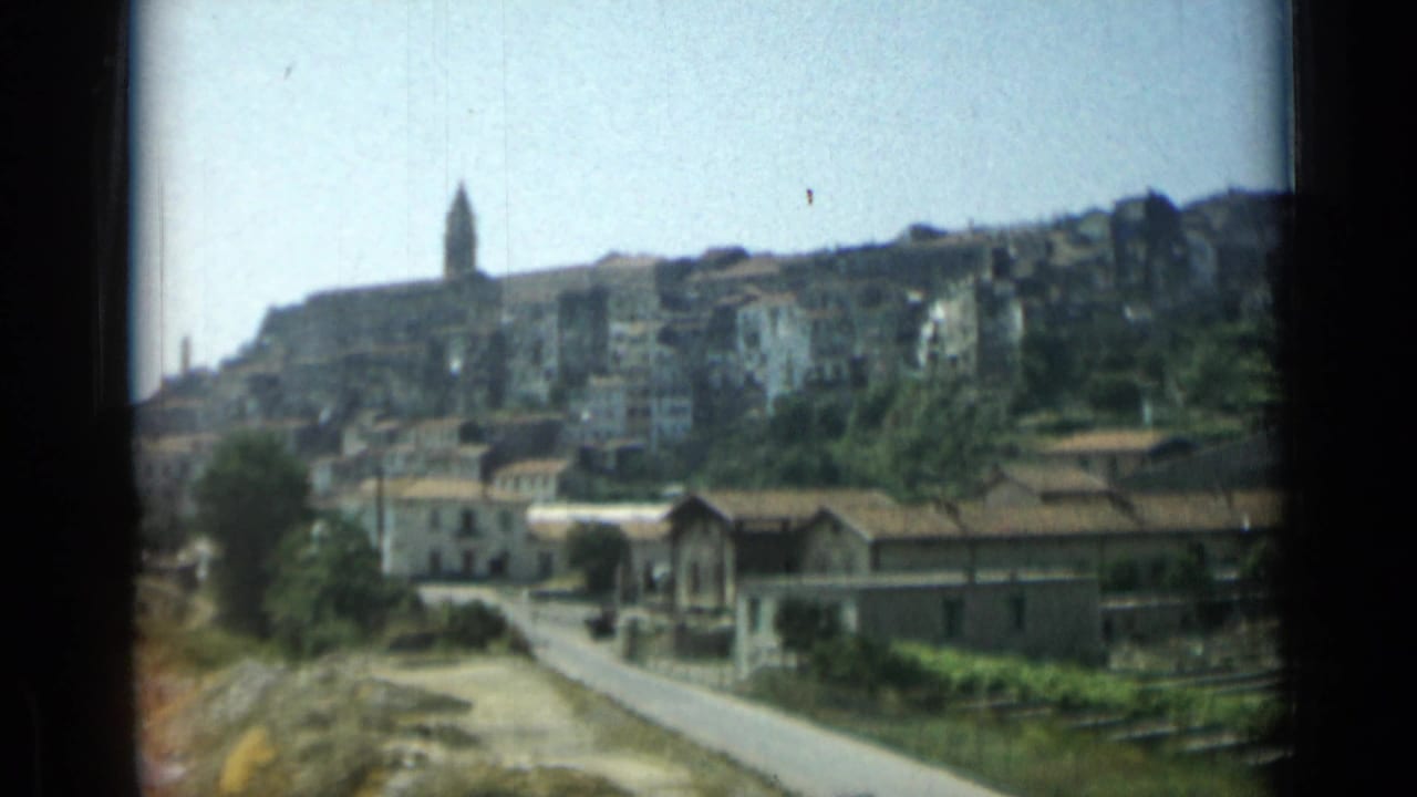 Hillside town with a church steeple above a road and foreground buildings.