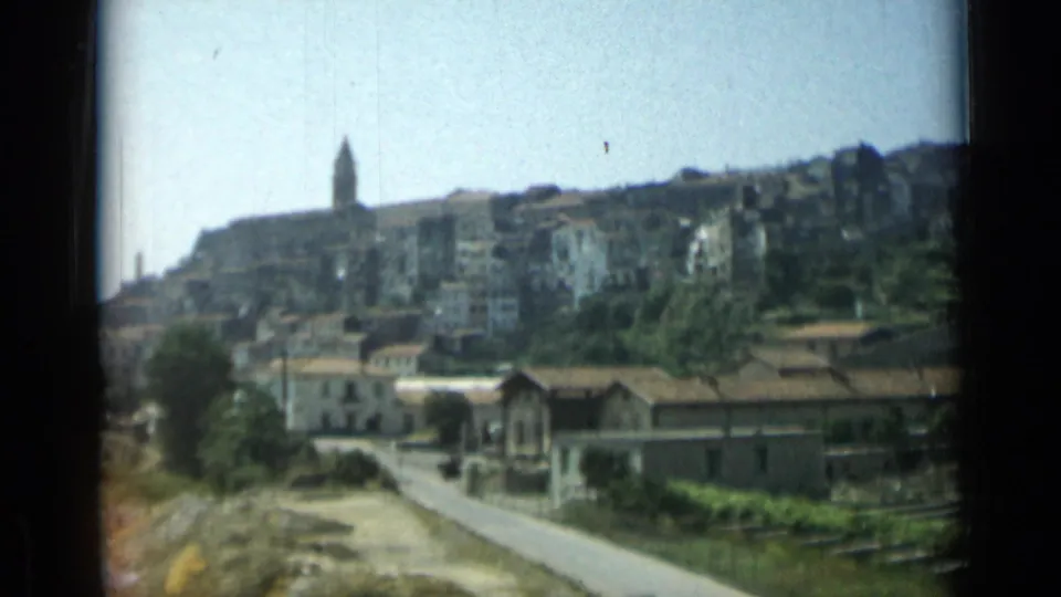 1962 Monaco home-movie frame showing a hillside town, church steeple, road, trees, and low buildings in bright daylight.