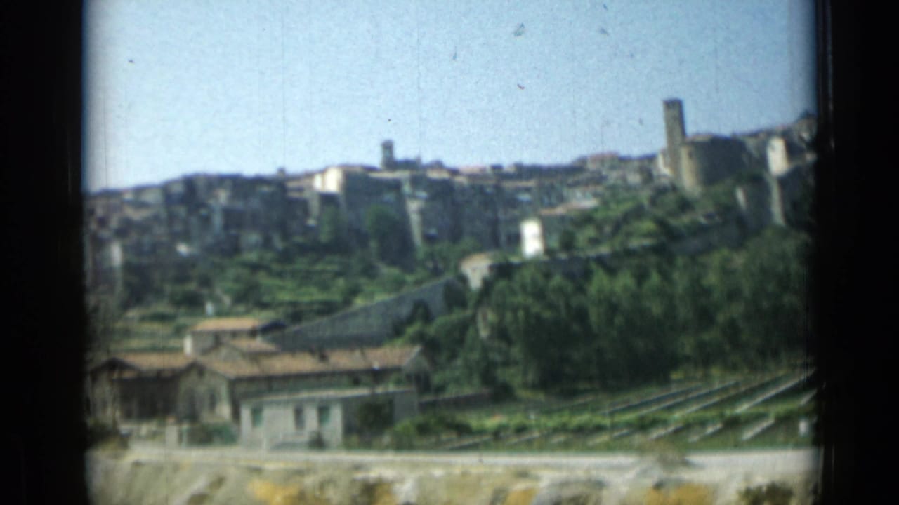 Hillside town with stone buildings, greenery, terraced plots, and low houses in the foreground.