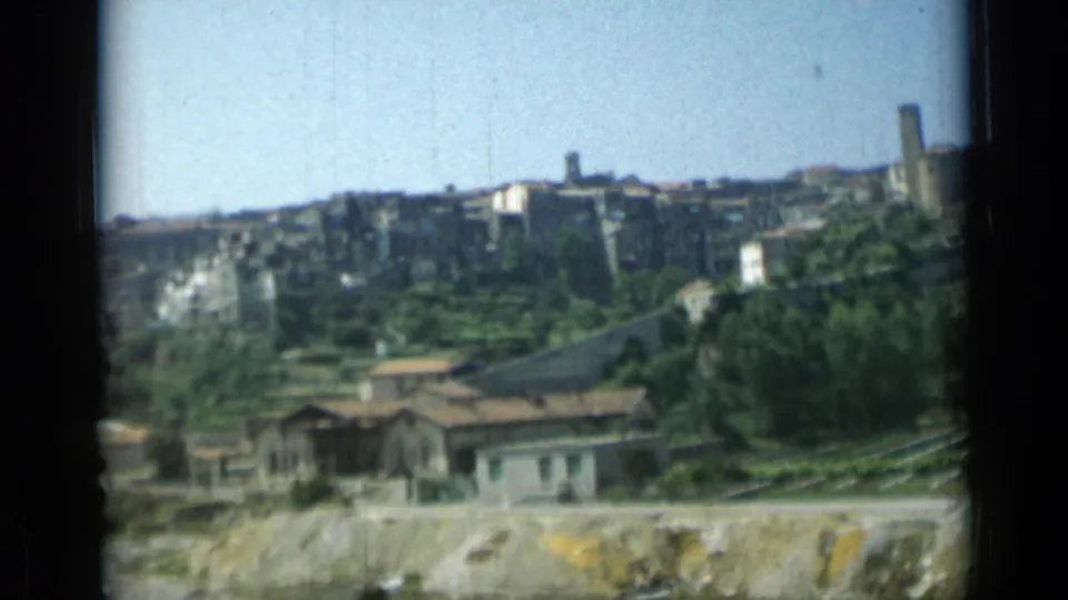 1962 home-movie frame showing a hillside settlement above terraced greenery and low structures near visible lines below.