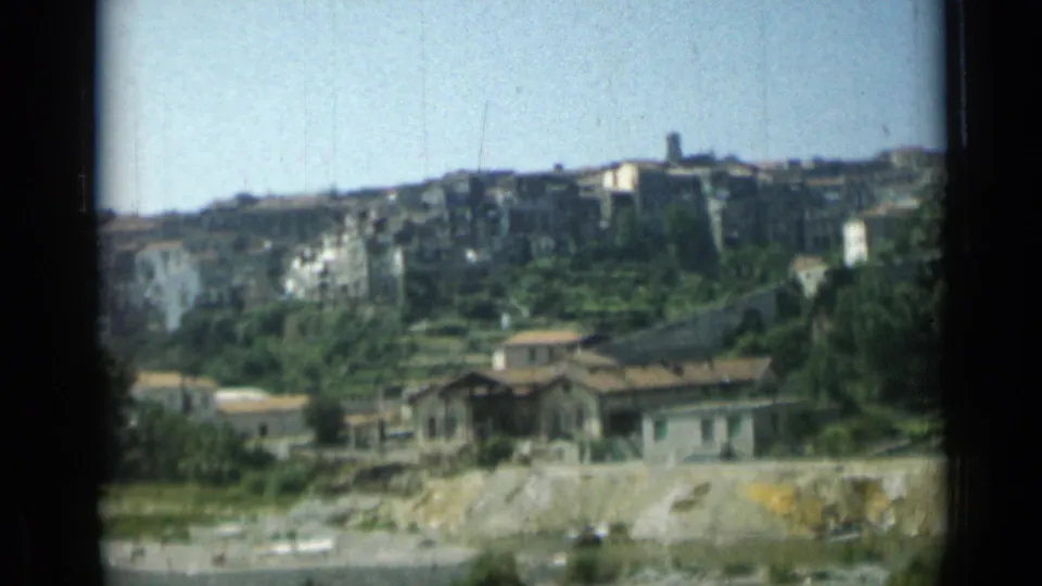 Soft 1962 8mm frame of a hillside town, with stone buildings above terraced plantings and low houses in front.