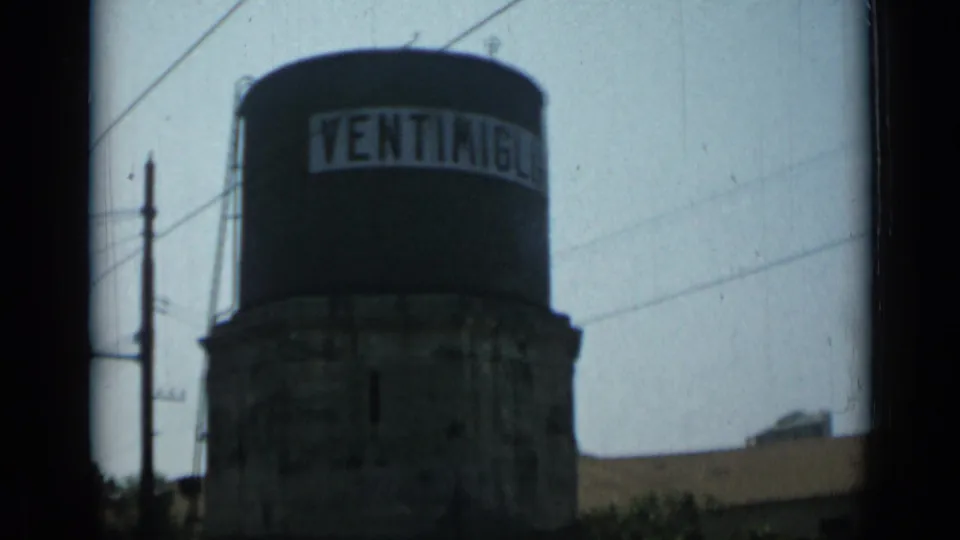 1962 8mm frame of a water tower labeled Ventimiglia, with utility wires and low surrounding structures visible.
