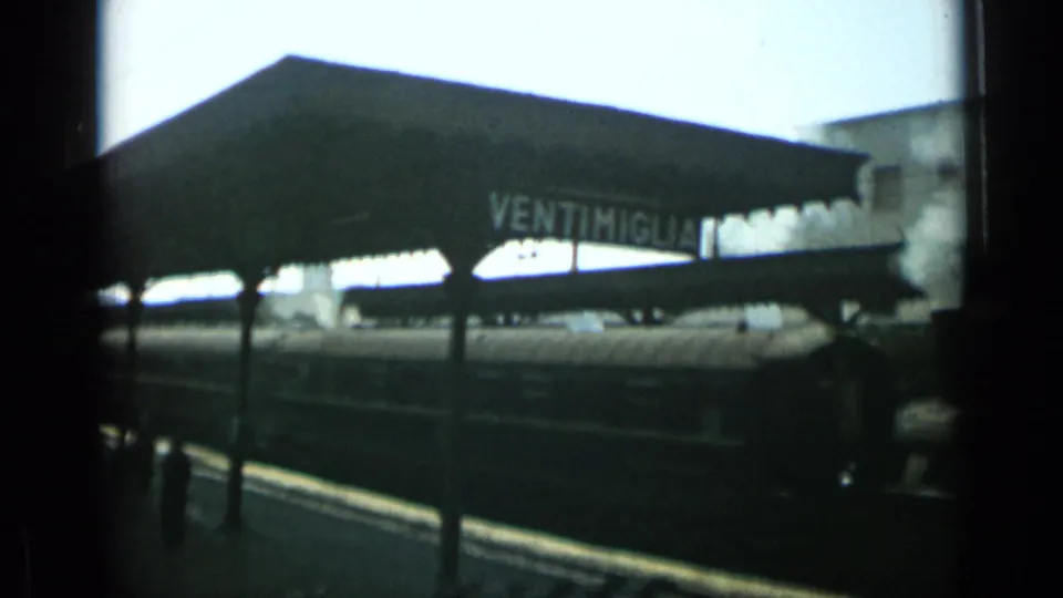 1962 8mm frame showing the Ventimiglia station name on a canopy with a train and platform in soft, dark exposure.