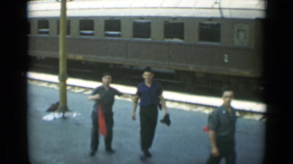 The group moves closer to the camera on the station platform, with the passenger car and platform post behind them.