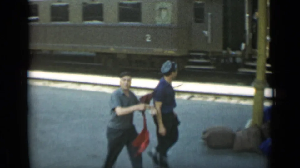 Two adult men pass nearer the camera on the Monaco platform, with bags or bundled items visible near the train.