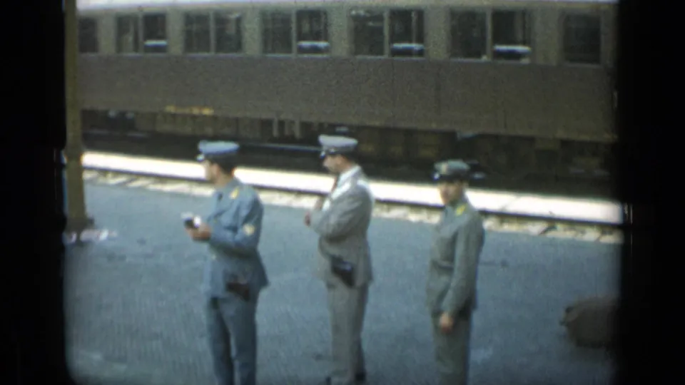 Three uniformed station officers stand apart on a Monaco rail platform in 1962, with a stationary passenger carriage behind them.