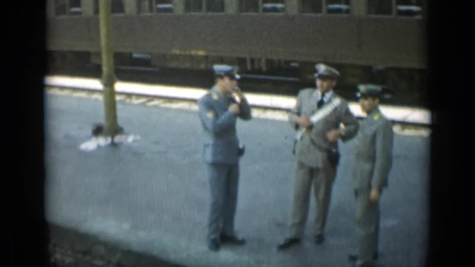 The home-movie camera frames three uniformed men at the Monaco station, with one smoking and another partly cut off at the edge.