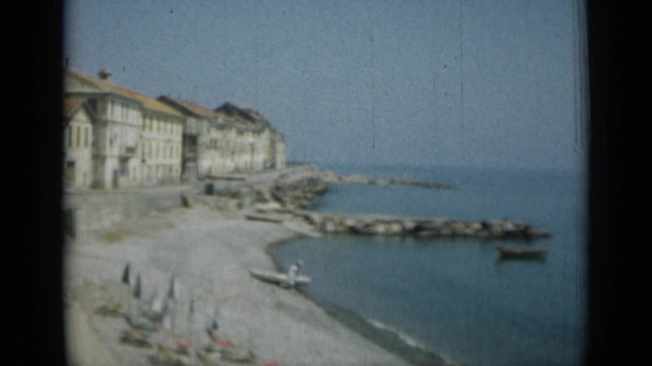 Waterfront buildings beside a pebble beach and blue sea with breakwater rocks.