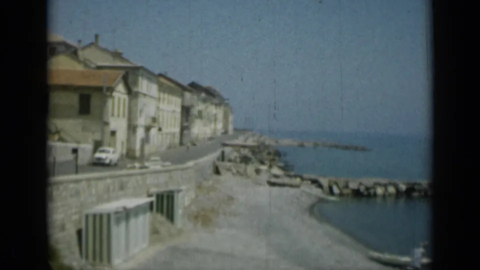 Monaco seaside road in 1962, with tan waterfront buildings, a white car, seawall, rocky breakwater, and blue water.
