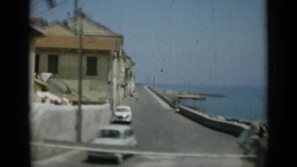 Passing 8mm view of a Monaco waterfront road with parked cars, a tan building, utility pole, seawall, and sea beyond.
