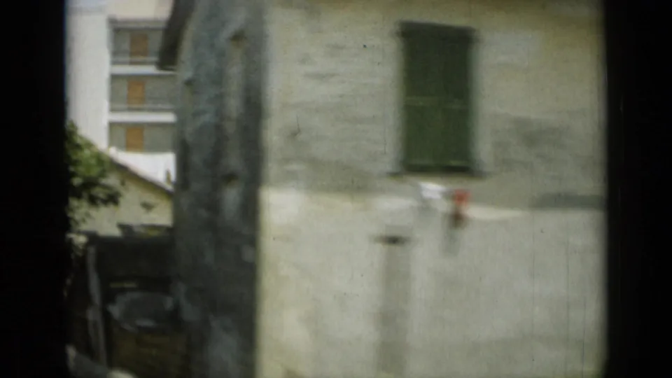 1962 Monaco home-movie view past a pale stone building with green shutters, nearby trees, and a dark vertical window-frame edge.