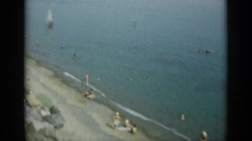 1962 Monaco coastal frame showing swimmers near a rocky shore and a small sailboat on calm blue water.