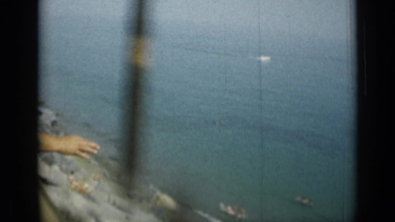 Rocky shoreline and blue sea seen through dark film-frame edges, with a partial arm at the left edge.