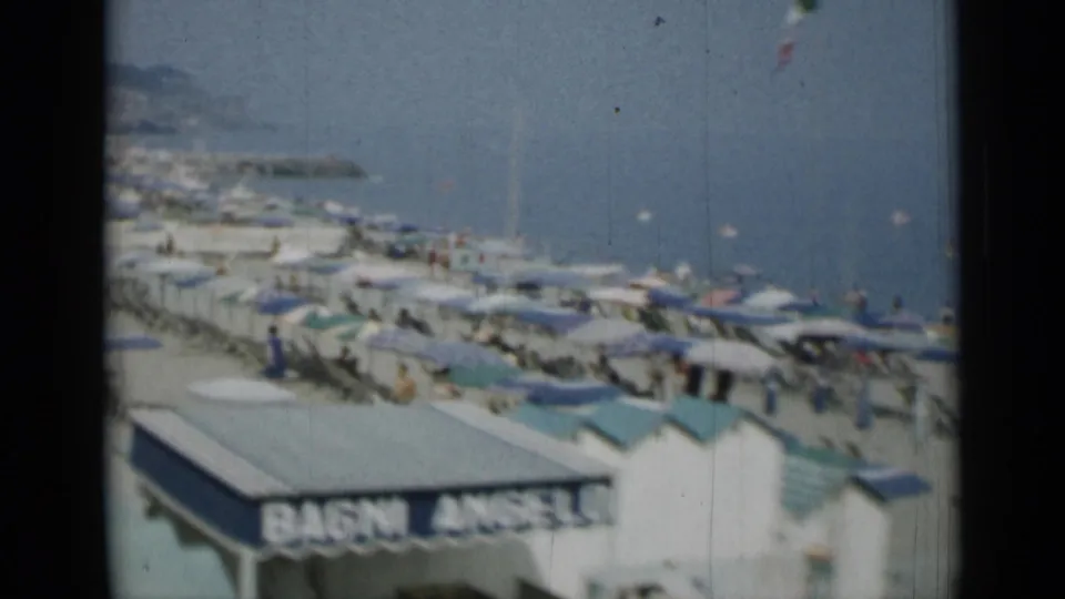 1962 8mm resort beach view in Monaco or nearby Italy, showing rows of umbrellas, beachgoers, and a hazy coastline.