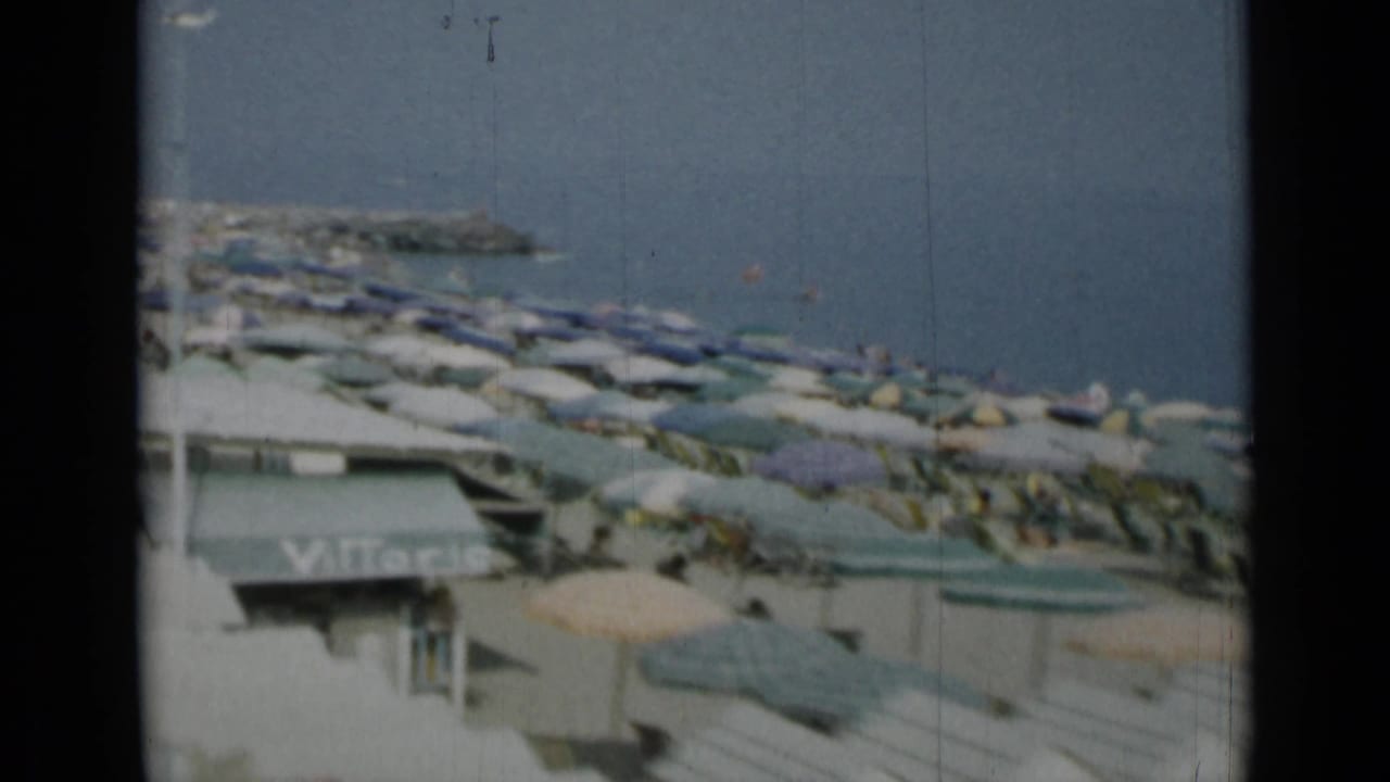Crowded beach with open umbrellas, lounge chairs, sea and a distant rocky breakwater.