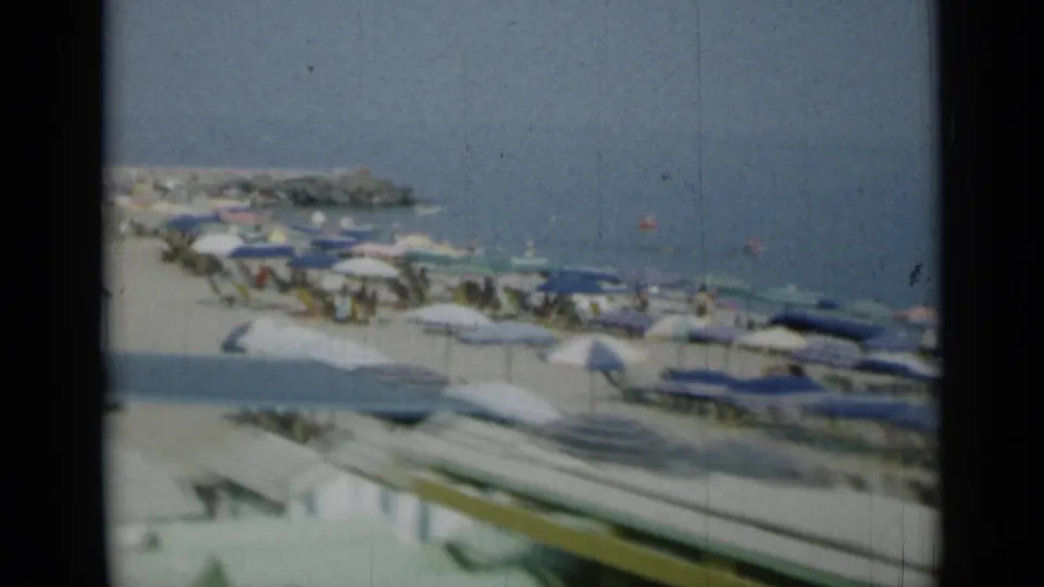 1962 Monaco home-movie frame of a busy beach terrace with umbrellas and pale cabanas near the sea.