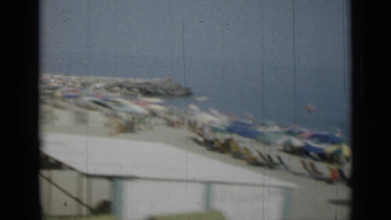 Soft 8mm view of a beach with umbrellas, boats and a rocky breakwater beyond a dark frame edge.