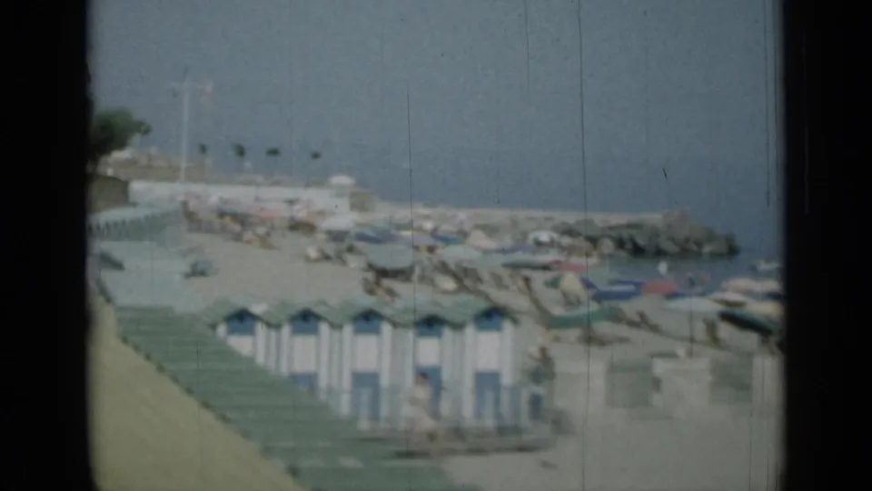 1962 Monaco seaside frame with blue-and-white beach huts, boats, a breakwater and calm water, partially blocked by the film framing.