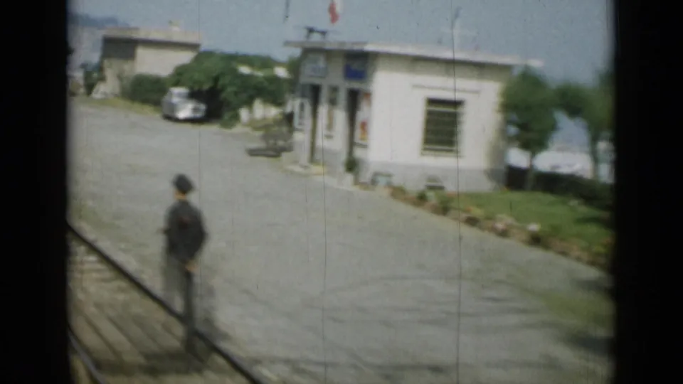 1962 Monaco coastal frame showing a seaside building, paved walkway, railings, shrubs and a distant standing person.