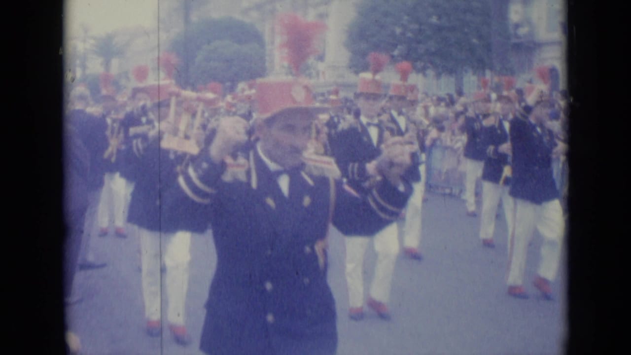 Marching band members in dark uniforms and red plumed hats play brass instruments in a street parade.