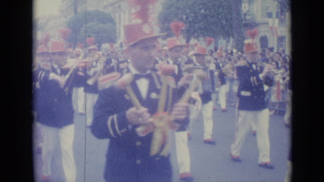 Uniformed marching band members in red hats parade along a city street.