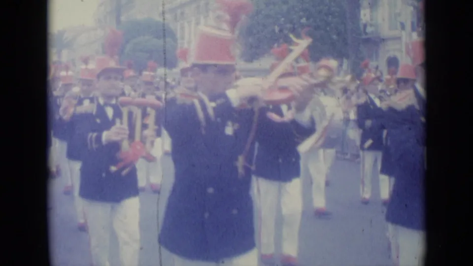 A Nice parade frame shows a uniformed musician in a red hat playing among other band members on a city street.