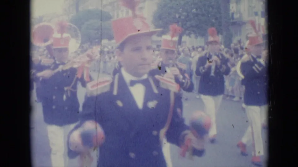 Final Nice parade frame with a red-hatted band member in front and brass musicians continuing behind on the street.