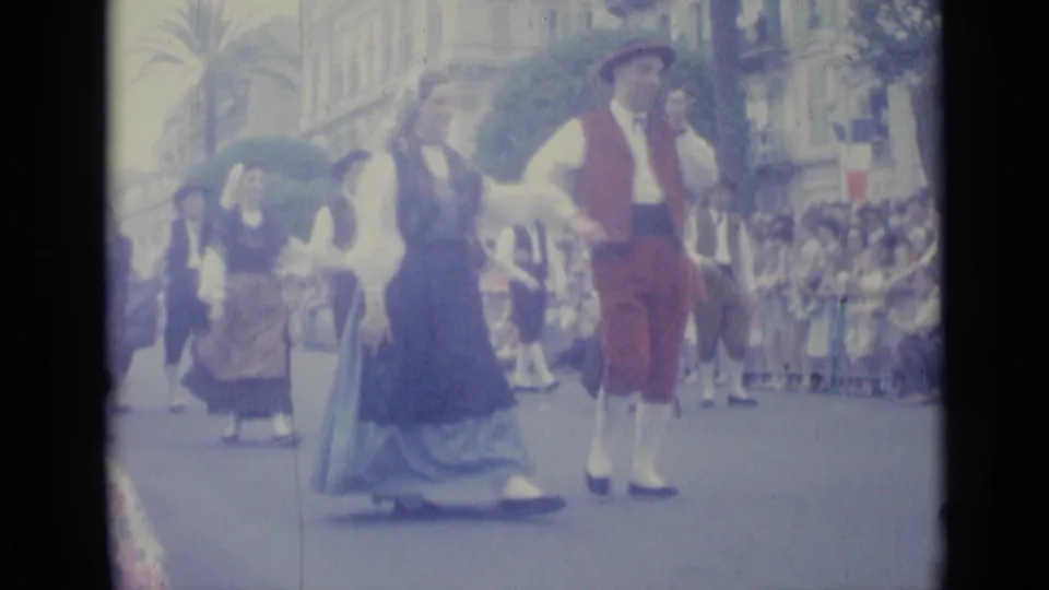 1962 Nice, France home-movie frame showing costumed men and women parading past curbside spectators on a city street.