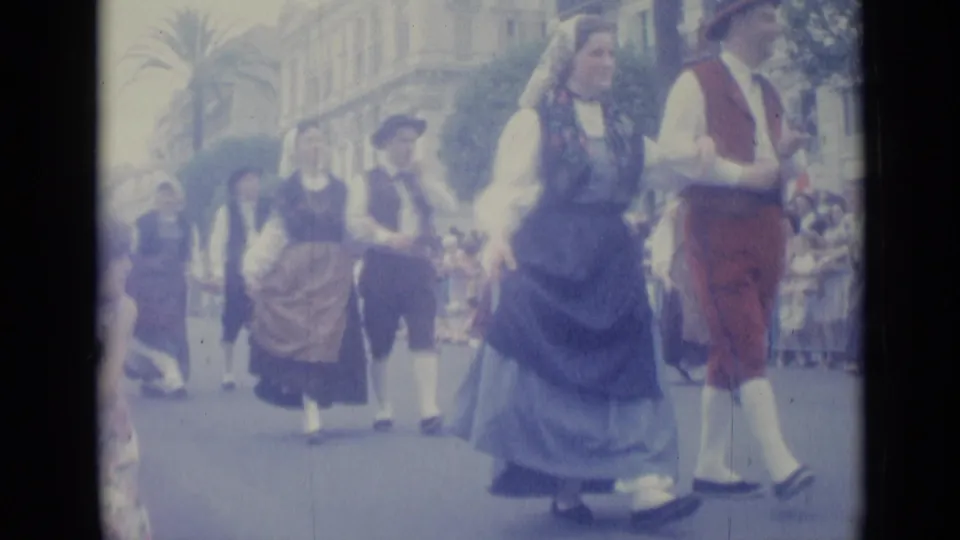 Costumed couple in a 1962 Nice street parade moves past the camera, with other participants and a watching crowd behind.