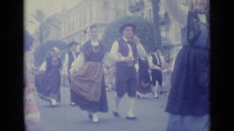 1962 Nice parade frame with a woman in a long skirt and a man in a vest walking together before other costumed participants.
