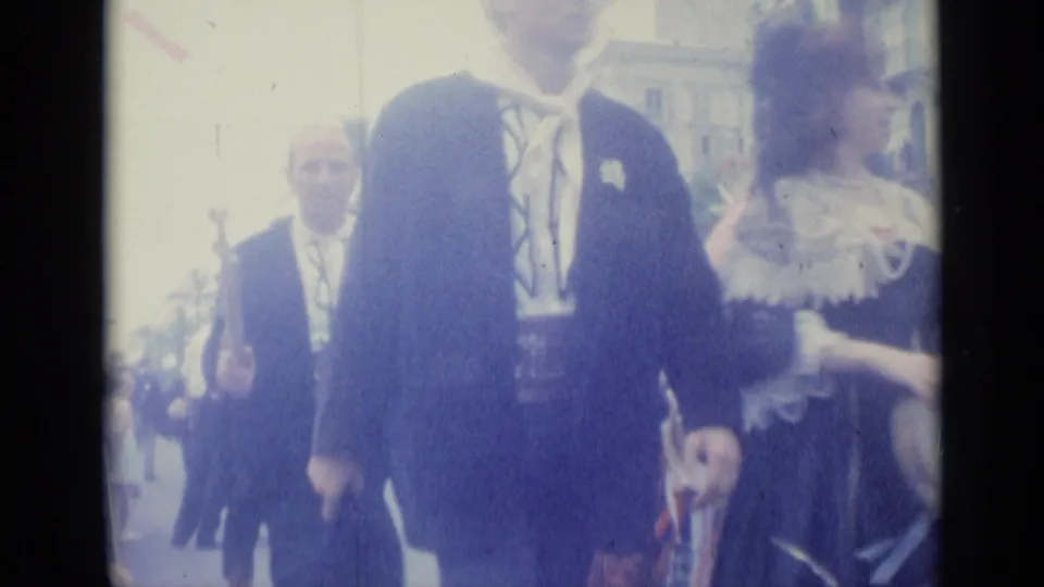 A close handheld view shows men and a woman in traditional attire moving through a Nice street procession in 1962.