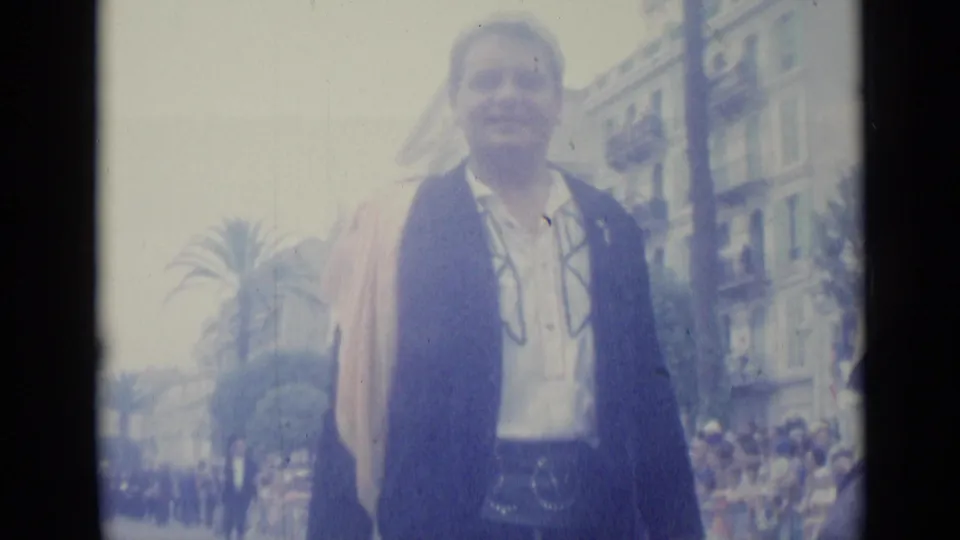A smiling procession participant in dark jacket, white shirt, sash, and belt stands on a Nice street in 1962.