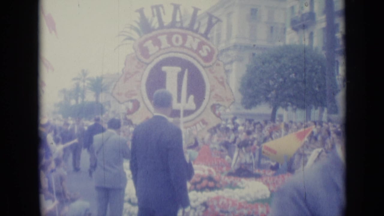 Parade float with a large Lions emblem, suited men, flowers, crowd, palm trees, and city buildings.