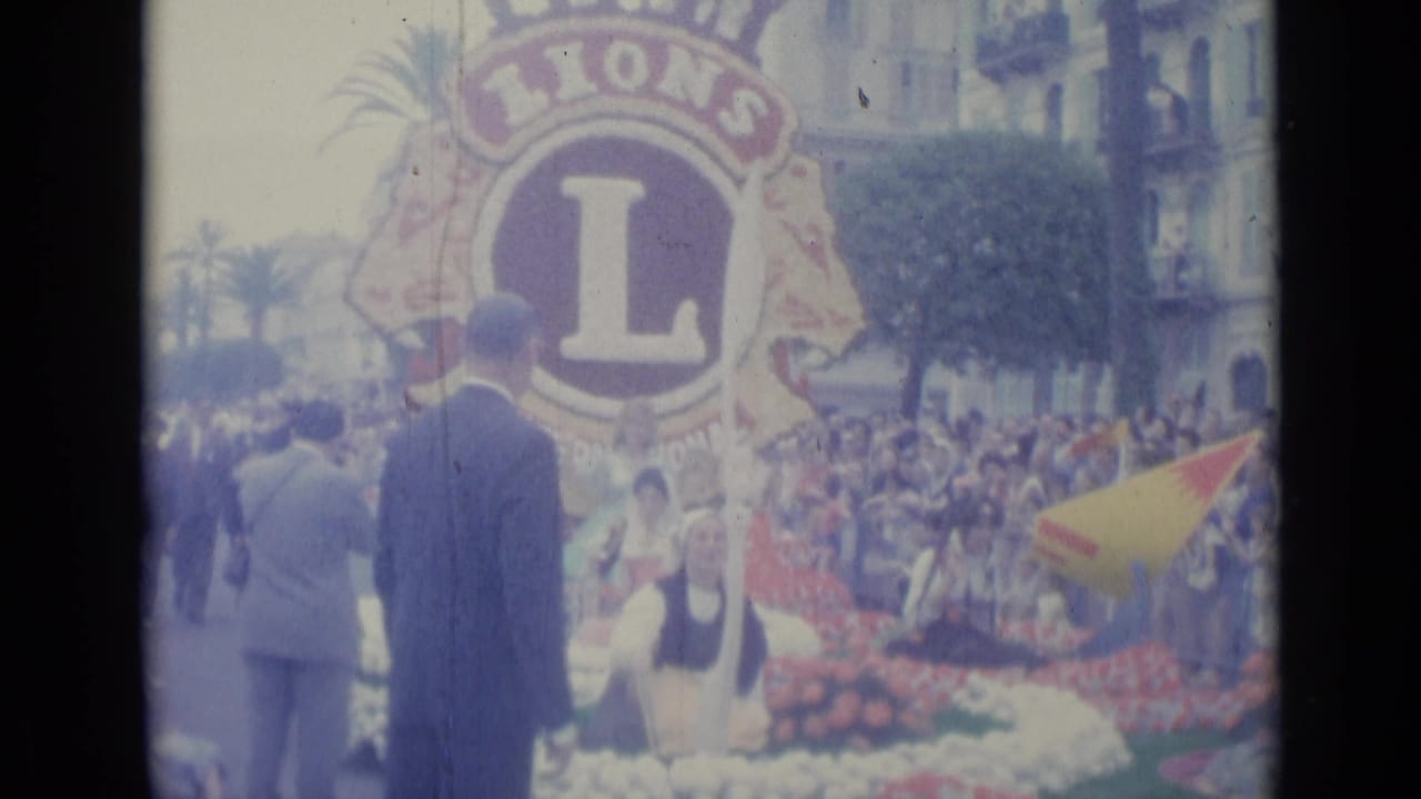 Large Lions emblem on a flower-covered float with spectators, suited men, palm trees, and buildings behind.