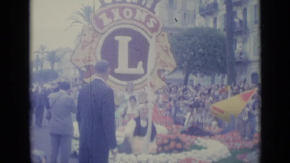 The Lions International emblem dominates a decorated parade float in Nice, with suited men and a dense crowd visible around it.