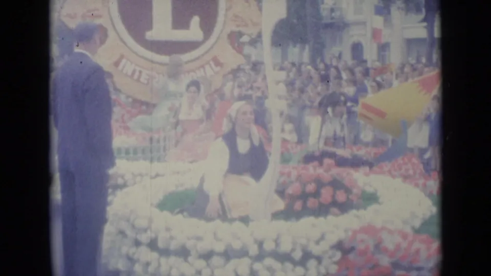 On the Nice parade float, a woman in traditional dress sits among flowers below a large Lions emblem as spectators watch nearby.