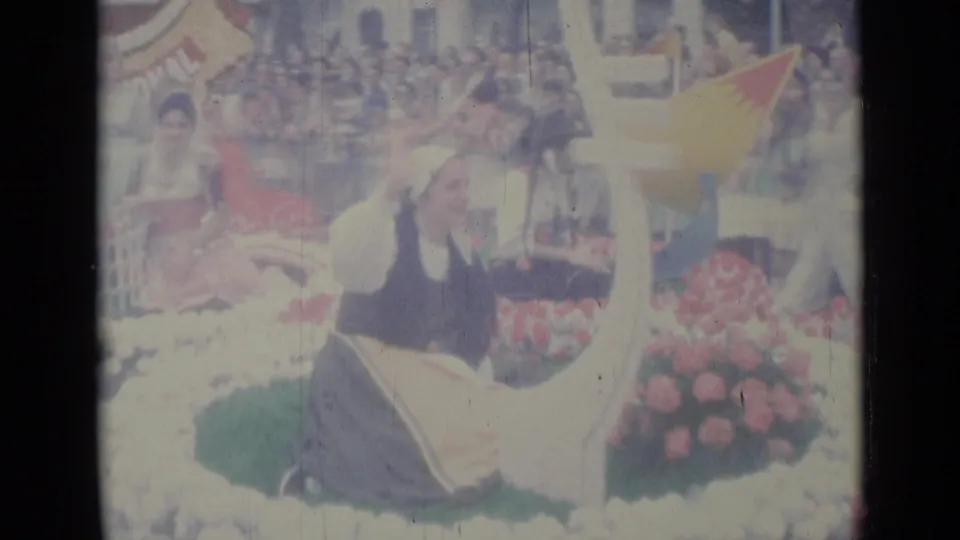 A costumed participant sits on the flower-covered Lions parade float in Nice, with a yellow pennant, emblem edge, and spectators behind.