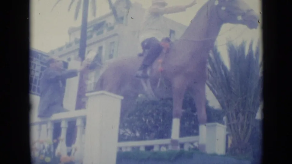 A brown horse and light-uniformed rider pass a white railing and blue timing board in Nice, France, in 1962 home-movie footage.