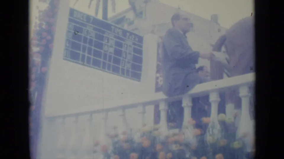 A suited spectator sits on the white railing as the horse passes near the blue timing board in 1962 Nice, France footage.