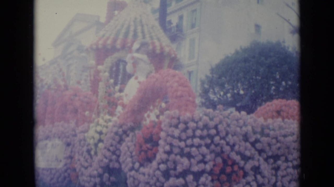 Woman in white on a flower-covered parade float with spectators and buildings behind her.