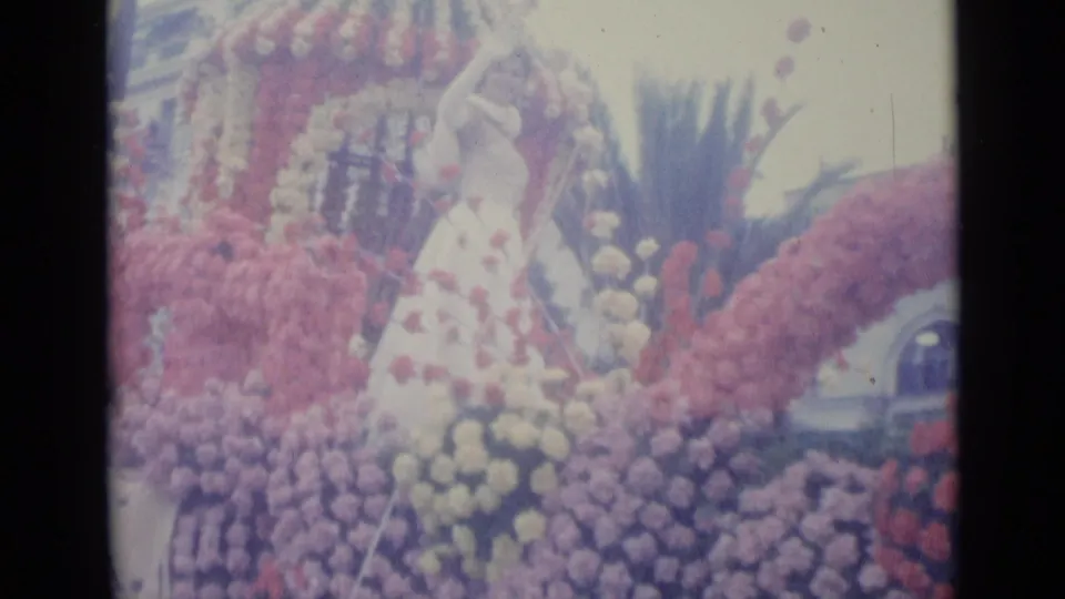 A close home-movie view shows the woman on the Nice parade float framed by floral arches, dense blossoms, and a palm-like plant behind her.