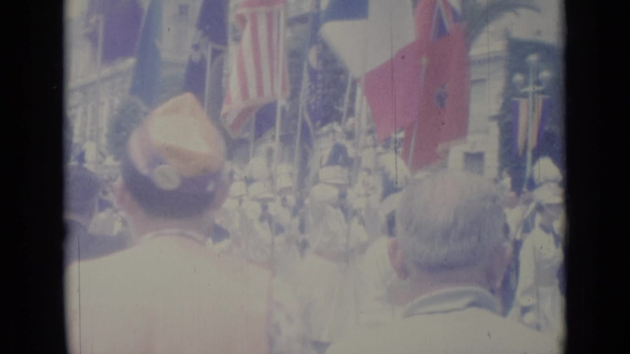 Crowd watches a parade with flags and uniformed marchers in the street.