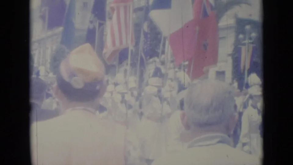 1962 Nice home-movie frame showing spectators in the foreground as flags and uniformed marchers pass along a city street.